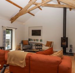 A living room with exposed wooden beams an orange sofa armchairs a wood stove and a bookshelf at 3 Caddaford Barns in Buckfastleigh