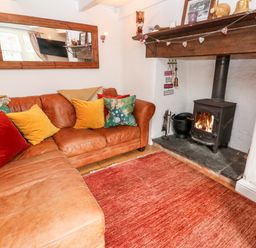 A living room with a leather sofa, colorful cushions, a wood-burning stove, and a large wall mirror at Honeysuckle Cottage in St Agnes