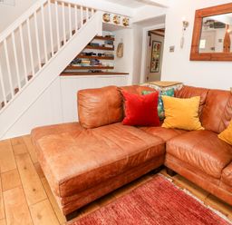 A living room with a brown leather sectional sofa with colorful cushions under a white staircase with shelves at Honeysuckle Cottage in St Agnes
