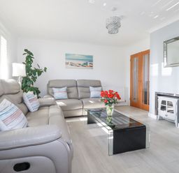 A living room with gray sofas a glass coffee table with red flowers a plant a white lamp a painting wooden doors and a white fireplace at Palm View in Holywell Bay