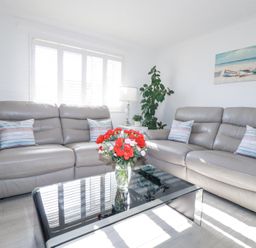 A living room with two leather sofas a glass coffee table with a vase of flowers and a painting of boats on the wall at Palm View in Holywell Bay