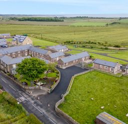 A cluster of stone buildings with pitched roofs surrounded by green fields and a road with slow markings at Glebe Cottage in Embleton