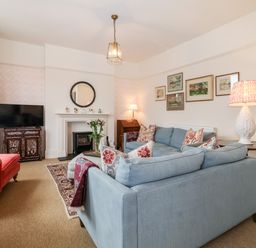 A living room with a blue sectional sofa red armchair television on cabinet fireplace and framed paintings at Collett House in Southwold