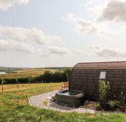 A curved roof cabin with a hot tub and potted plants in a field with rolling hills and a lake in the background at Ash in Saltash