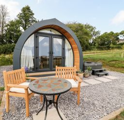 An outdoor seating area with wooden chairs and a mosaic table in front of a modern pod-style cabin with glass doors and a hot tub at Ash in Saltash