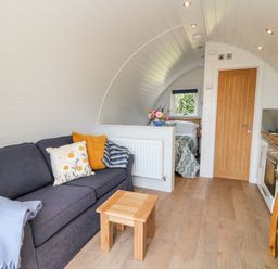 A living area with a dark sofa and cushions a small wooden table kitchenette with stools a door and a bed in a small room at Ash in Saltash