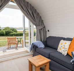 A living area with a dark sofa and cushions next to an open door leading to an outdoor seating area with wooden chairs and a table at Ash in Saltash