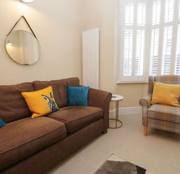 A living room with a brown sofa and cushions a patterned armchair and a round mirror at Derwent Cottage in Keswick