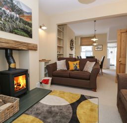 A living room with a wood burning stove a brown sofa with cushions and a dining area visible in the background at Derwent Cottage in Keswick