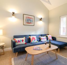 A living room with a blue sofa and colorful cushions a wooden coffee table with wine glasses and a bottle a side table with a lamp and window shutters at Poppy Cottage Southwold