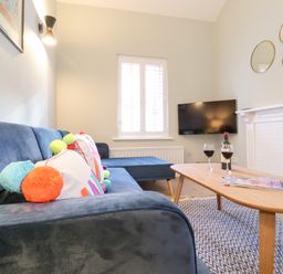 A living room with a blue sofa and colorful pillows a wooden coffee table with two glasses of red wine a bottle and magazines a wall mounted TV and three round mirrors above a white fireplace at Poppy Cottage in Southwold
