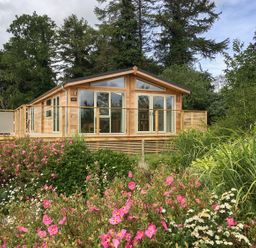 A wooden cabin with glass doors surrounded by trees and flowering bushes at 2 Faraway Fields in Dobwalls