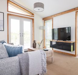 A living room with a sofa and cushions a coffee table with vases a wall mounted television and large windows at 6 Southern Place in Dobwalls