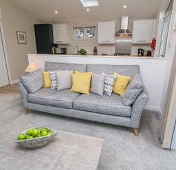 A living room with a grey sofa and yellow and grey pillows a coffee table with green apples and a kitchen in background at 4 Faraway Fields Dobwalls