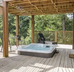 An outdoor deck with a hot tub two chairs a small table and wooden railing surrounded by trees at 4 Faraway Fields in Dobwalls