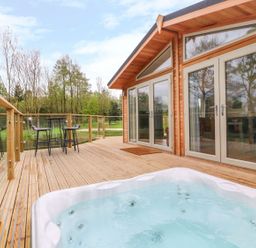 A wooden deck with a hot tub and outdoor seating next to a cabin with glass doors at 21 Meadow Retreat in Dobwalls