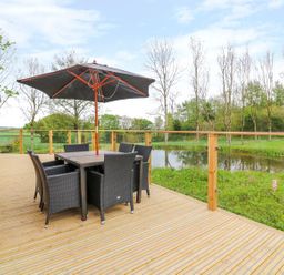 A wooden deck with a dining table set and umbrella overlooking a pond and trees at 21 Meadow Retreat in Dobwalls