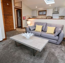 A living room with a gray sofa with yellow cushions a rectangular coffee table and kitchen area in the background at 2 Southern Place in Dobwalls