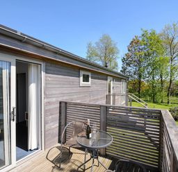 A balcony with a glass table and two chairs outside a wooden house with trees in the background at 19 Meadow Retreat in Dobwalls