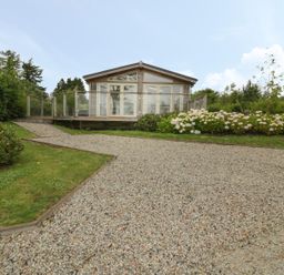 A gravel driveway leading to a small house with glass doors and a wooden deck surrounded by bushes and flowers at 11 Faraway Fields in Dobwalls