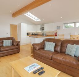 A living room with two brown leather sofas a wooden coffee table with remotes and a kitchen with white cabinets and a wooden breakfast bar at 11 Faraway Fields in Dobwalls