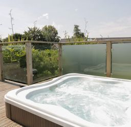 An outdoor hot tub on a wooden deck with glass panel railing and plants behind at 12 Faraway Fields in Dobwalls
