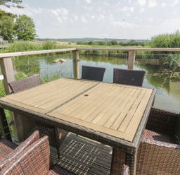 An outdoor deck with a wooden table and six wicker chairs overlooking a pond at 15 Faraway Fields in Dobwalls