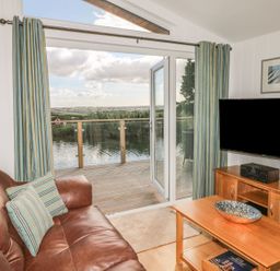 A living room with leather sofa and striped curtains opening to a deck overlooking water at 14 Faraway Fields in Dobwalls