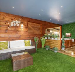 An indoor room with artificial grass flooring a wicker sofa with cushions a wicker table wooden walls and a wooden deck with a spa bath and various potted plants at 9 Meadow Retreat in Dobwalls