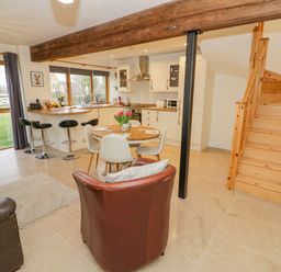 An open plan room with a kitchen, dining table, sofa, armchair, bar stools, and wooden staircase at The Granary in Little Dasset near Gaydon