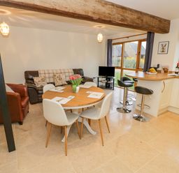 An open plan living area with a round dining table and chairs a brown sofa a leather armchair bar stools at a kitchen counter and large windows showing a garden at The Granary Little Dasset near Gaydon