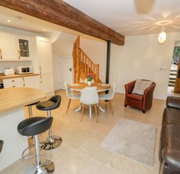 An open plan kitchen and living area with a round dining table and chairs a leather sofa an armchair bar stools and wooden stair at The Granary in Little Dasset near Gaydon