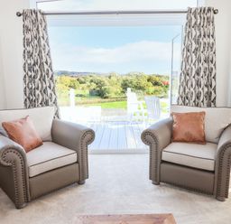 Two armchairs with cushions facing a large open window with a view of outdoor chairs and greenery at Chestnut Lodge in Teigngrace near Newton Abbot