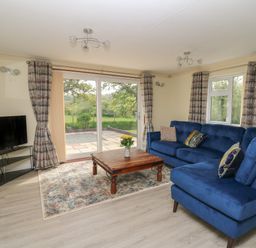 A living room with a blue sectional sofa a wooden coffee table a television on a stand and large windows with curtains at Erwlon in Rhydyfelin near Aberystwyth