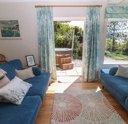 A sitting room with two blue sofas, patterned curtains, a rug, and a door opening to a garden with a hot tub at Fron Olau in Nefyn