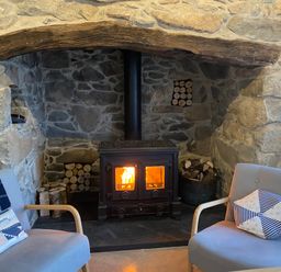A stone fireplace with a wood burning stove firewood stored inside and two armchairs with cushions in a living room at Fron Olau in Nefyn