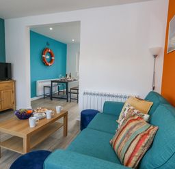 A living room with a blue sofa and colorful pillows a wooden coffee table with cups and fruit a TV on a wooden stand and a dining area with a life ring on the wall at Lagoon View in Wyke