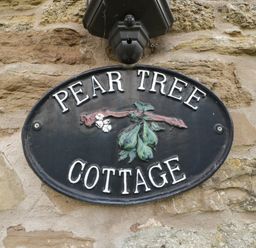 A black oval sign with pears hanging from a branch and the text Pear Tree Cottage on a stone wall at Peartree Cottage in Stanford Bishop near Bromyard