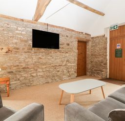 A living room with a stone wall a mounted television a wooden door a coffee table armchairs and a side table with a lamp at Peartree Cottage in Stanford Bishop near Bromyard