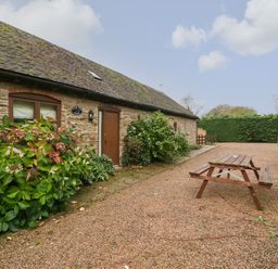 A stone cottage with a wooden door and window surrounded by plants and a picnic table on a gravel path at Clover Patch Cottage Stanford Bishop near Bromyard