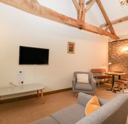 A living room with a wall-mounted television a gray armchair a gray sofa with a yellow cushion a dining table with chairs and a bench exposed wooden beams and a stone wall at Clover Patch Cottage in Stanford Bishop near Bromyard