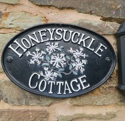 A black oval sign with white letters reading Honeysuckle Cottage mounted on a stone wall next to a lantern
