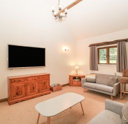A living room with a wall-mounted television a wooden cabinet a gray sofa a coffee table and a window with curtains at Honeysuckle Cottage in Stanford Bishop near Bromyard