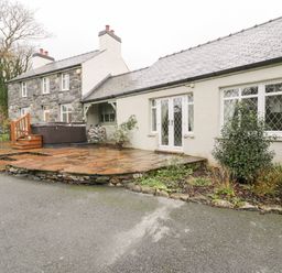 Exterior view of a stone and white painted house with a paved patio area hot tub and outdoor seating at Lakeside Cottage in Llanrug