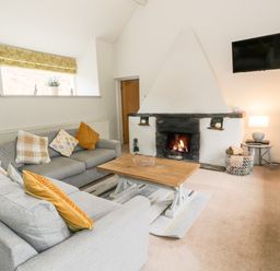A living room with two grey sofas cushions a wooden coffee table a fireplace with a fire a wall-mounted television a window with a yellow curtain and a side table with a lamp at Lakeside Cottage in Llanrug