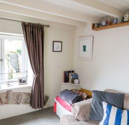 A corner room with a window with curtains a sofa with cushions a small shelf with books and framed pictures at 1 The Old Brewhouse in Dartmouth