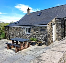 An outdoor patio area with a wooden picnic table and benches beside a stone house with a tiled roof and potted plants at Tir Eironwy in Dinas near Nefyn