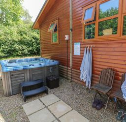 An outdoor area with a hot tub, two wooden chairs, towels hanging on hooks, and two pairs of sandals at 1 Hornbeam Lodge in Narberth