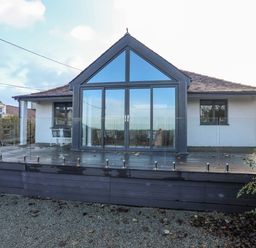 A house with large glass doors and windows facing a wooden deck with glass railing at Haulfryn in Moelfre