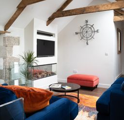 A living room with blue sofas orange cushions a black coffee table a small red ottoman a wall-mounted television a glass-enclosed fireplace a plant wooden beams and a compass wall decoration at The Rum House in Porthallow